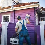 Main painting the outside of the house purple. Has a white bucket that reads "Kane painting"